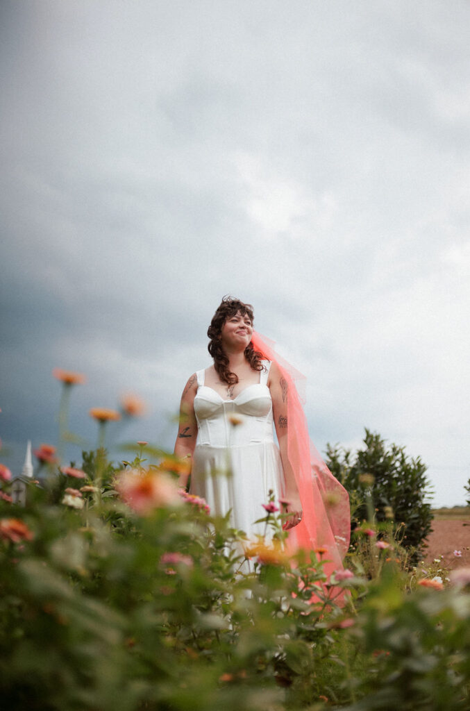 Bride with pink veil and white dress standing in wildflowers.