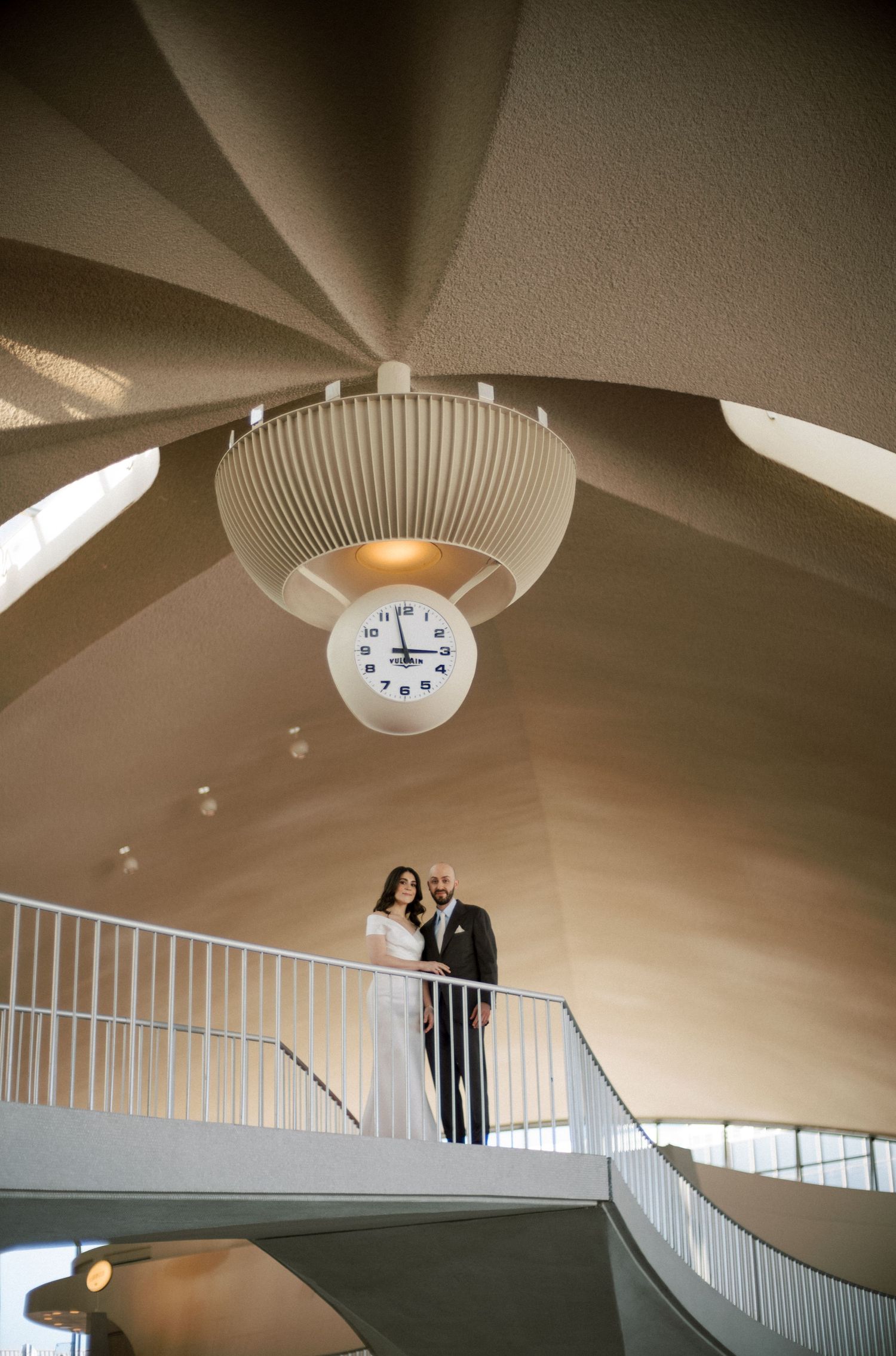 Bride and groom standing under clock at TWA Hotel wedding at JFK Airport.